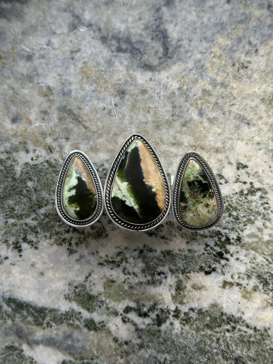 Three teardrop-shaped green and black gemstone rings with silver rope detailing on marble surface