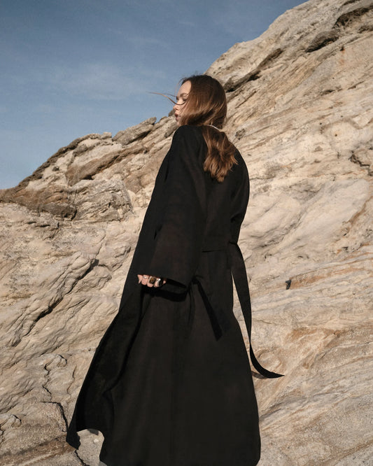 Woman in a long black coat standing against rocky cliff under clear blue sky