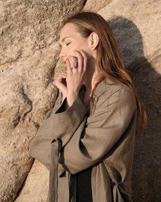 Woman wearing beige trench coat and large stone ring posing with eyes closed against rock background