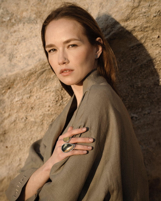 Portrait of a woman in a beige jacket wearing statement rings, posing against a textured rock wall