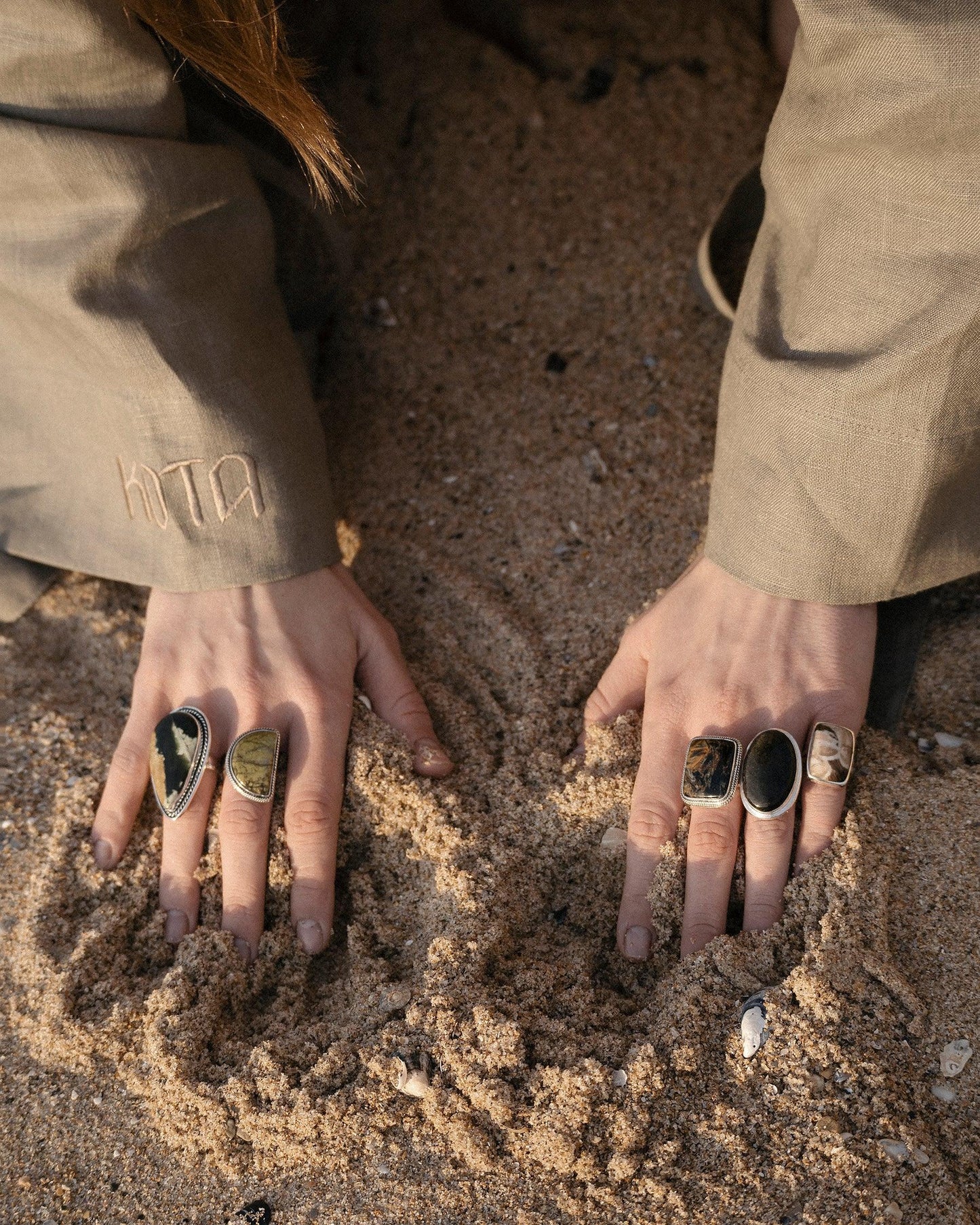 Hands with five unique statement rings, pressing into sandy beach, wearing beige sleeves embroidered with letters.