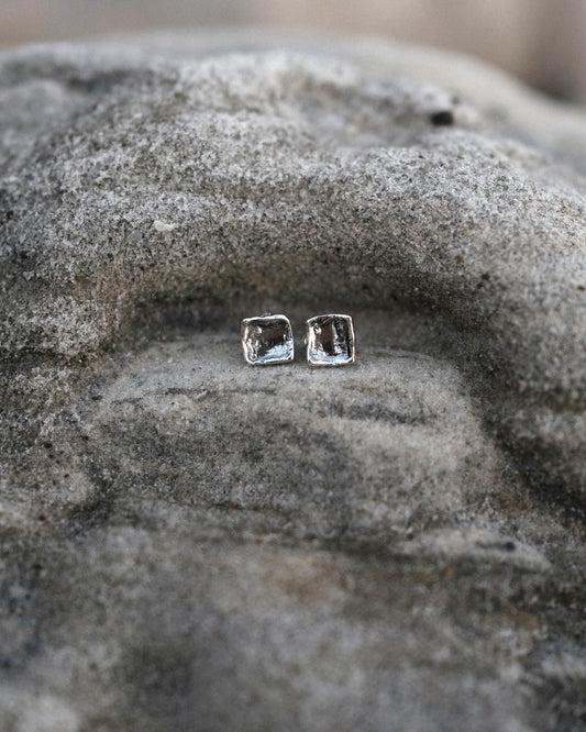 Minimalist silver square stud earrings on rough textured stone surface