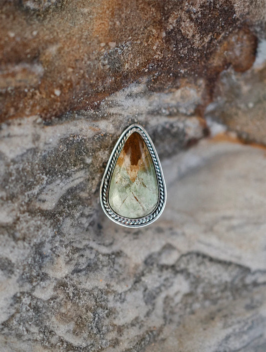 Teardrop-shaped pendant with green and brown gemstone, silver twisted frame on textured stone background