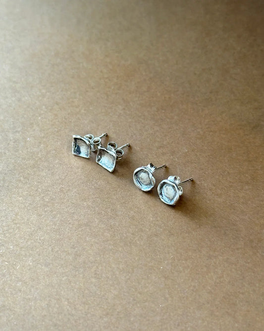 Two pairs of silver stud earrings with square and round shapes on brown surface