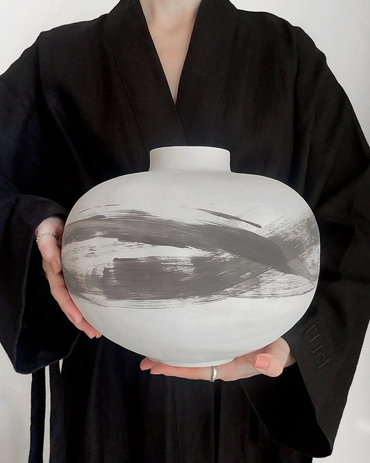 Person in black robe holding large white ceramic vase with gray brushstroke design