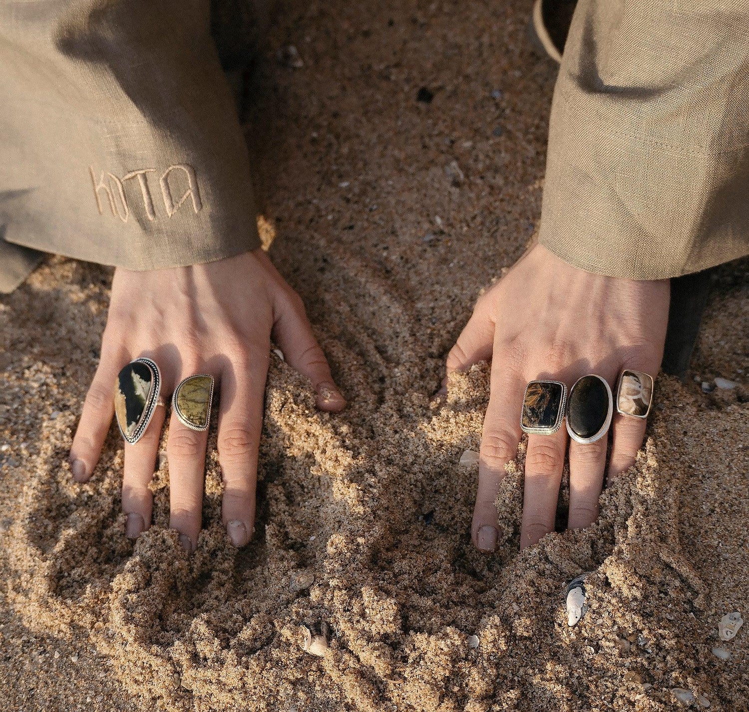 Close-up of hands with artistic stone rings partially buried in sandy beach with beige sleeves