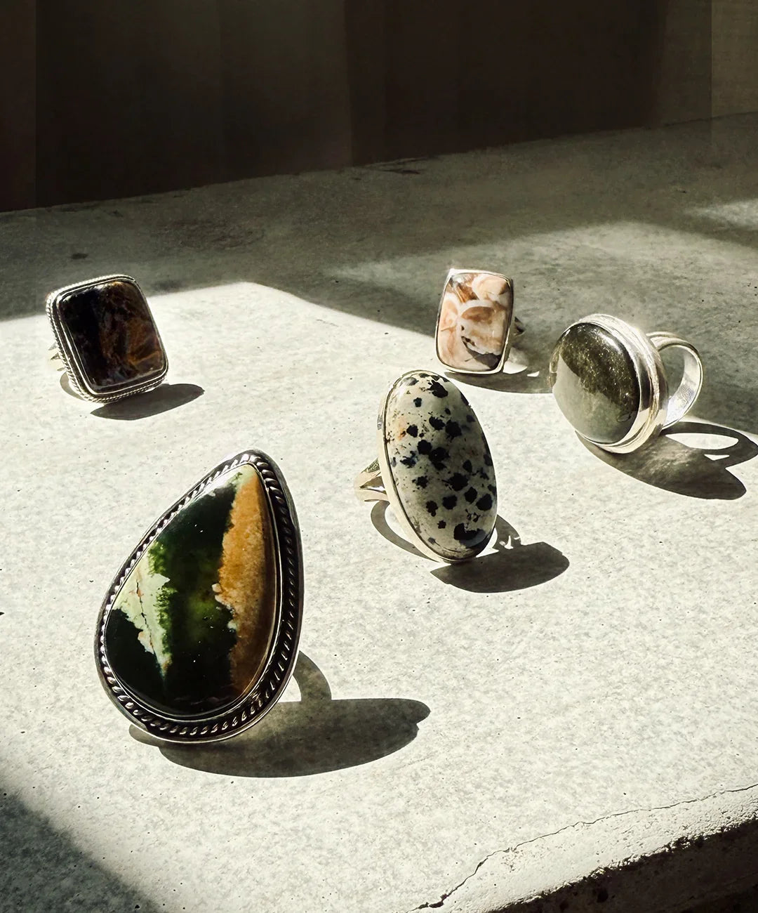 Five unique gemstone rings with silver settings displayed on a sunlit stone surface