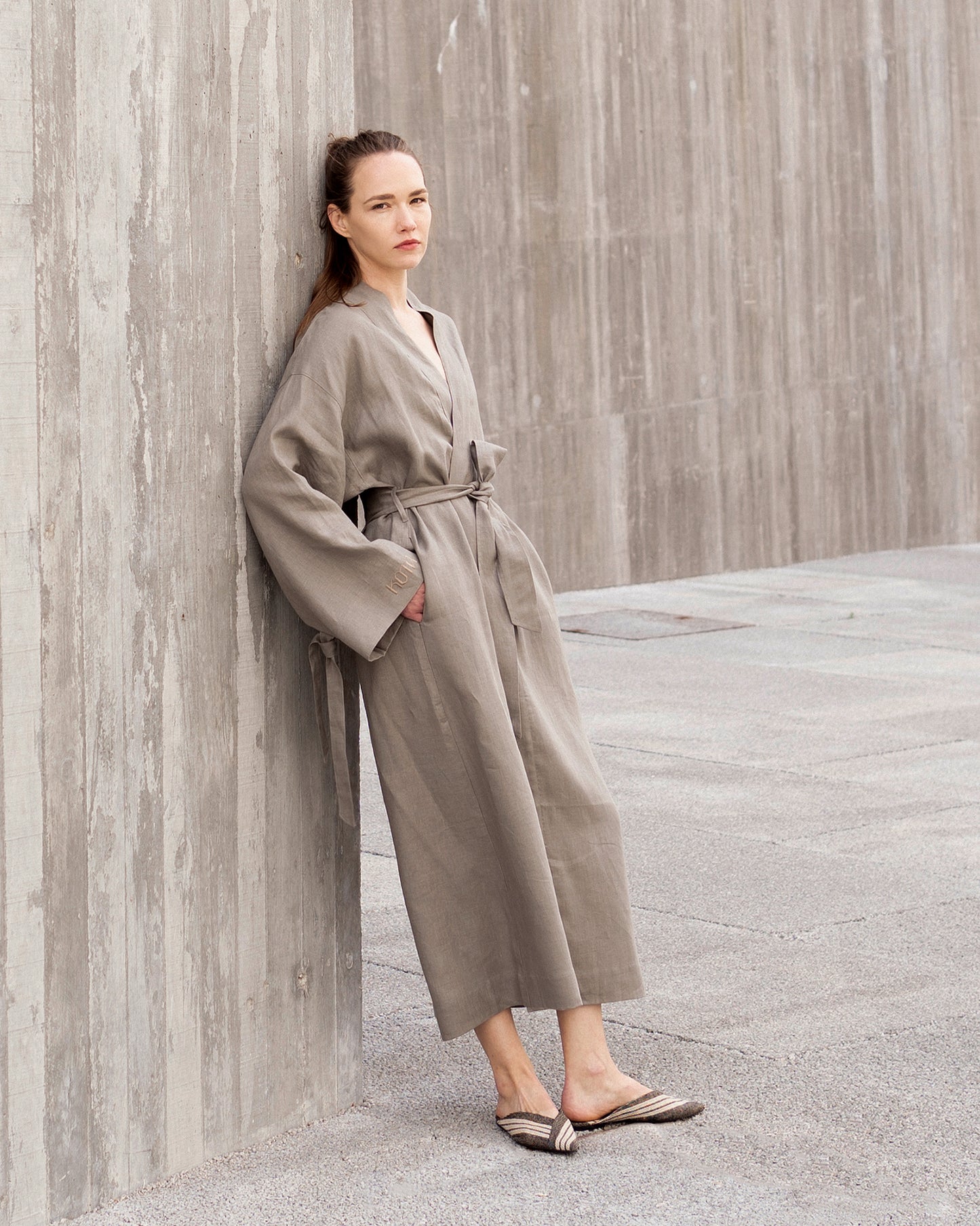 Woman in a grey robe standing against a textured wall.