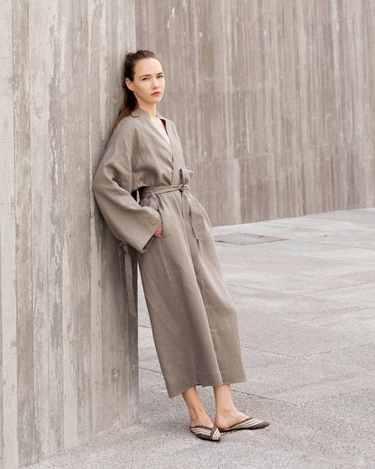 Woman in a grey robe standing against a textured wall.