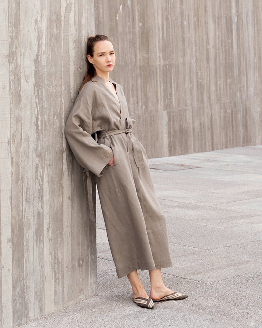 Woman in a grey robe standing against a textured wall.