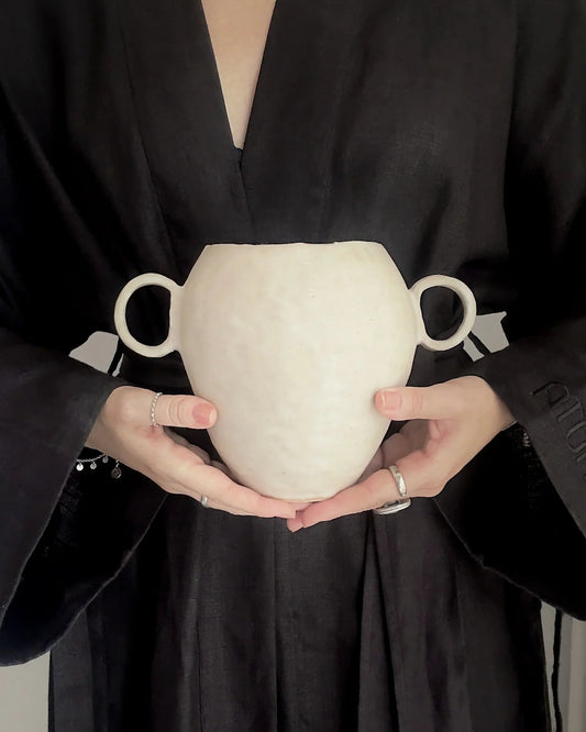 Person in black robe holding a white two-handled ceramic vase with silver rings