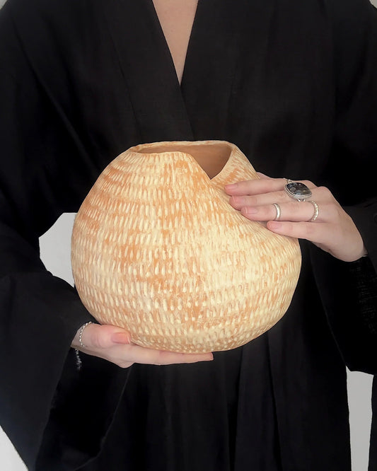 Hands holding a large textured beige ceramic vase with a fold, person wearing black robe