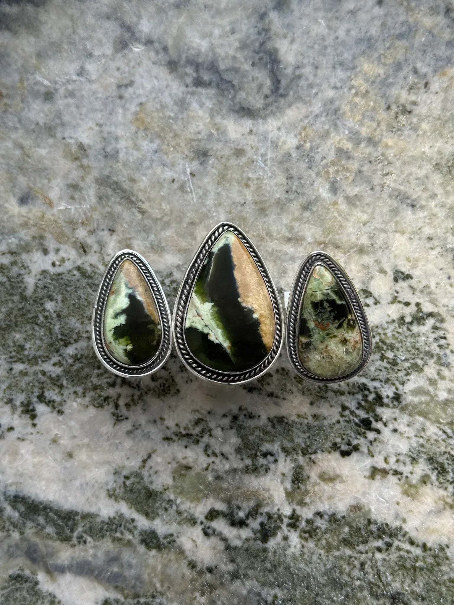 Three teardrop-shaped green and black gemstone rings with silver rope detailing on marble surface