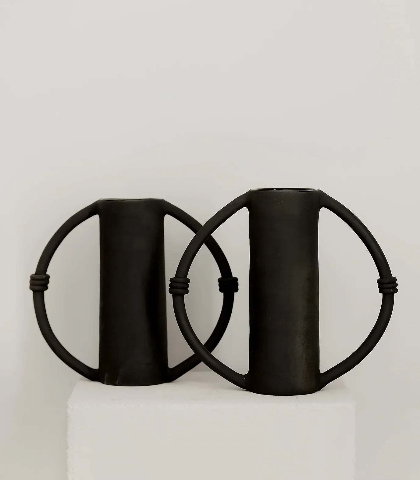 Two black handmade ceramic vases with circular side handles on a white pedestal