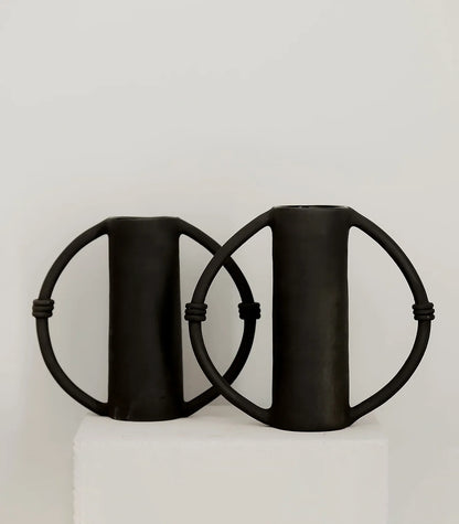 Two black handmade ceramic vases with circular side handles on a white pedestal