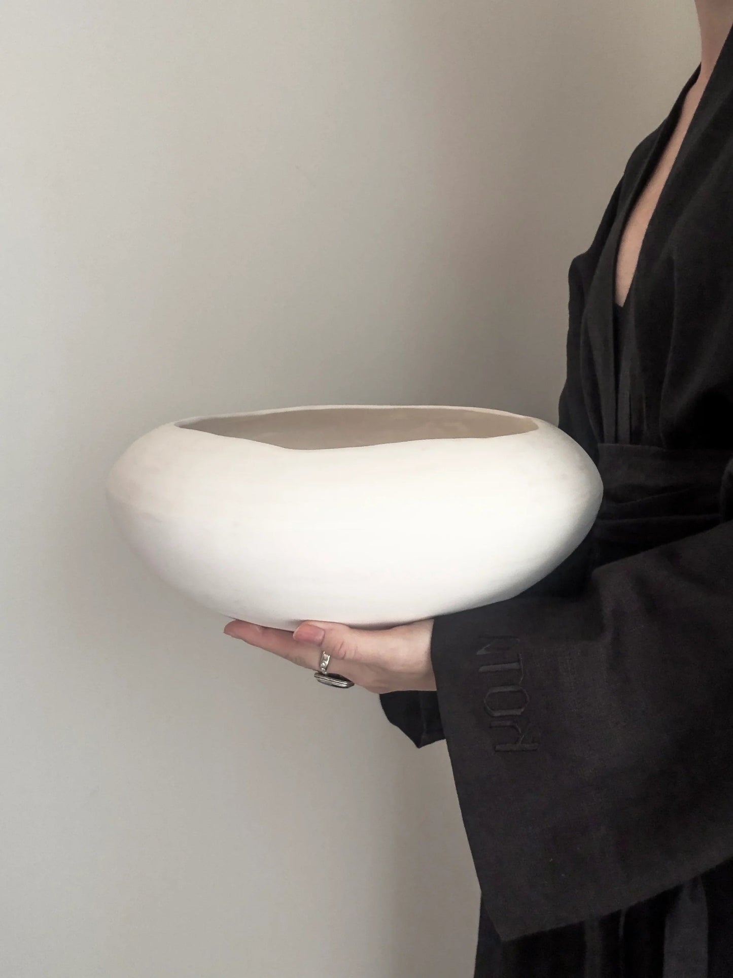 Person in black robe holding a large, white, minimalist ceramic bowl against a plain wall