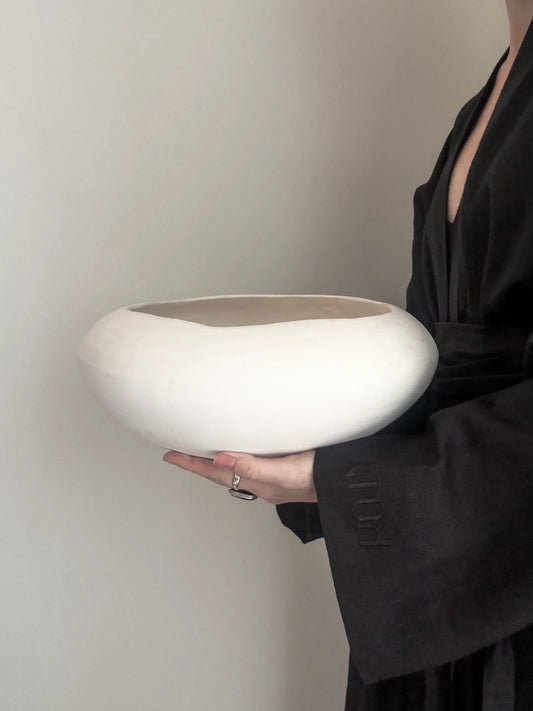 Person in black robe holding a large, white, minimalist ceramic bowl against a plain wall