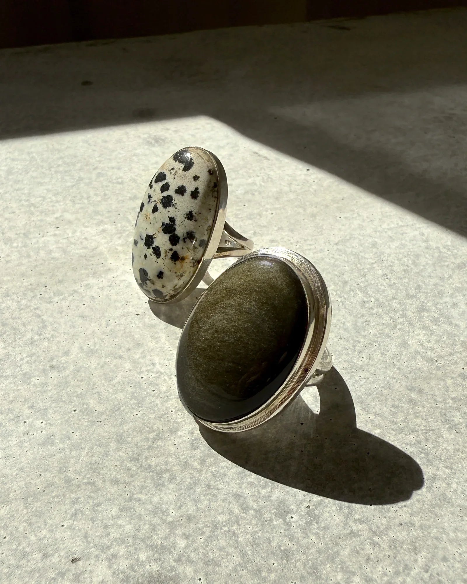 Two silver rings with large oval gemstones, one spotted black and white, the other dark green, on textured surface