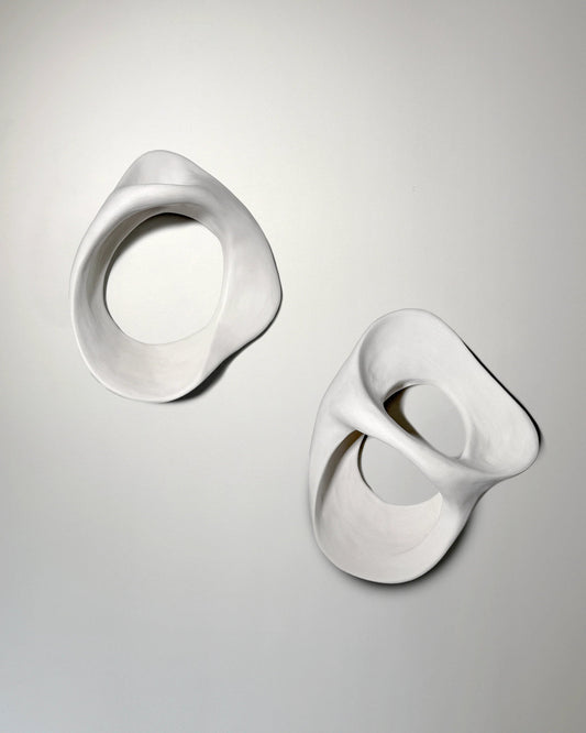Two white abstract ceramic sculptures with smooth, organic shapes mounted on a plain light wall