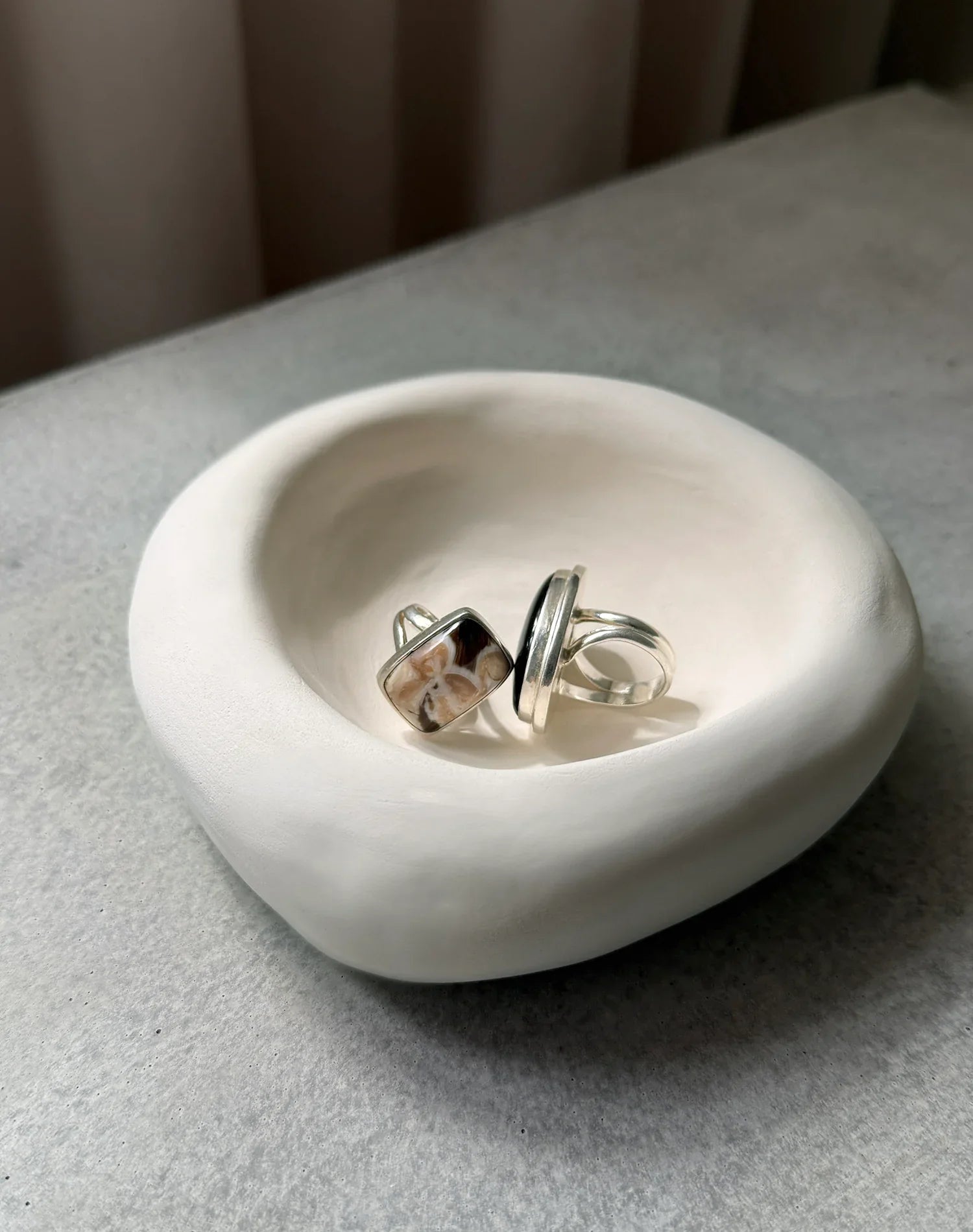 Two silver rings with polished stones in a smooth white ceramic bowl on a gray surface