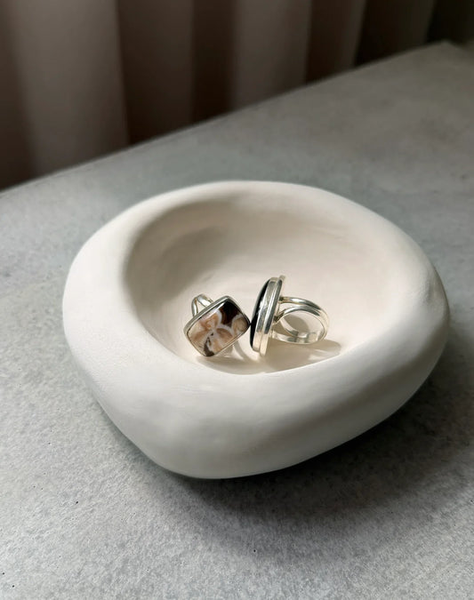 Two silver rings with polished stones in a smooth white ceramic bowl on a gray surface