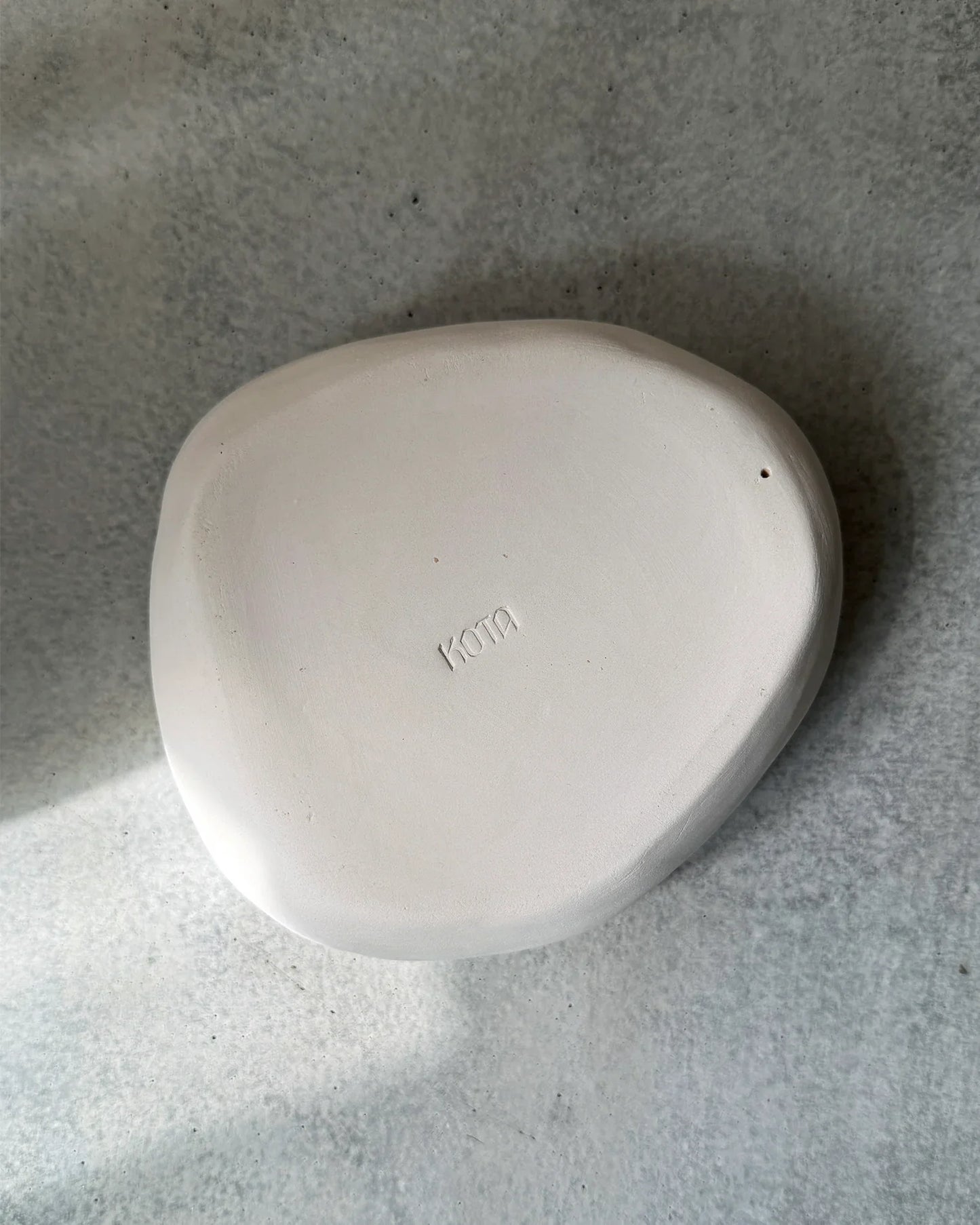 Minimalist curved white ceramic plate with 'Kota' inscription on textured grey surface