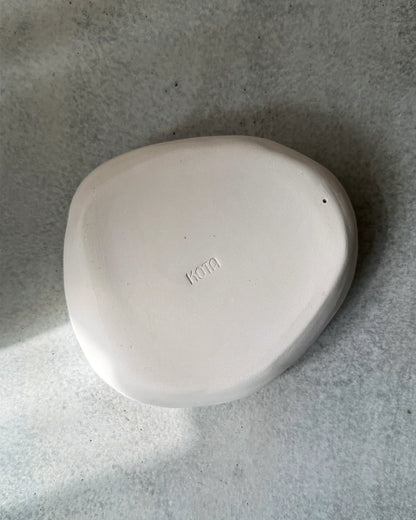 Minimalist curved white ceramic plate with 'Kota' inscription on textured grey surface