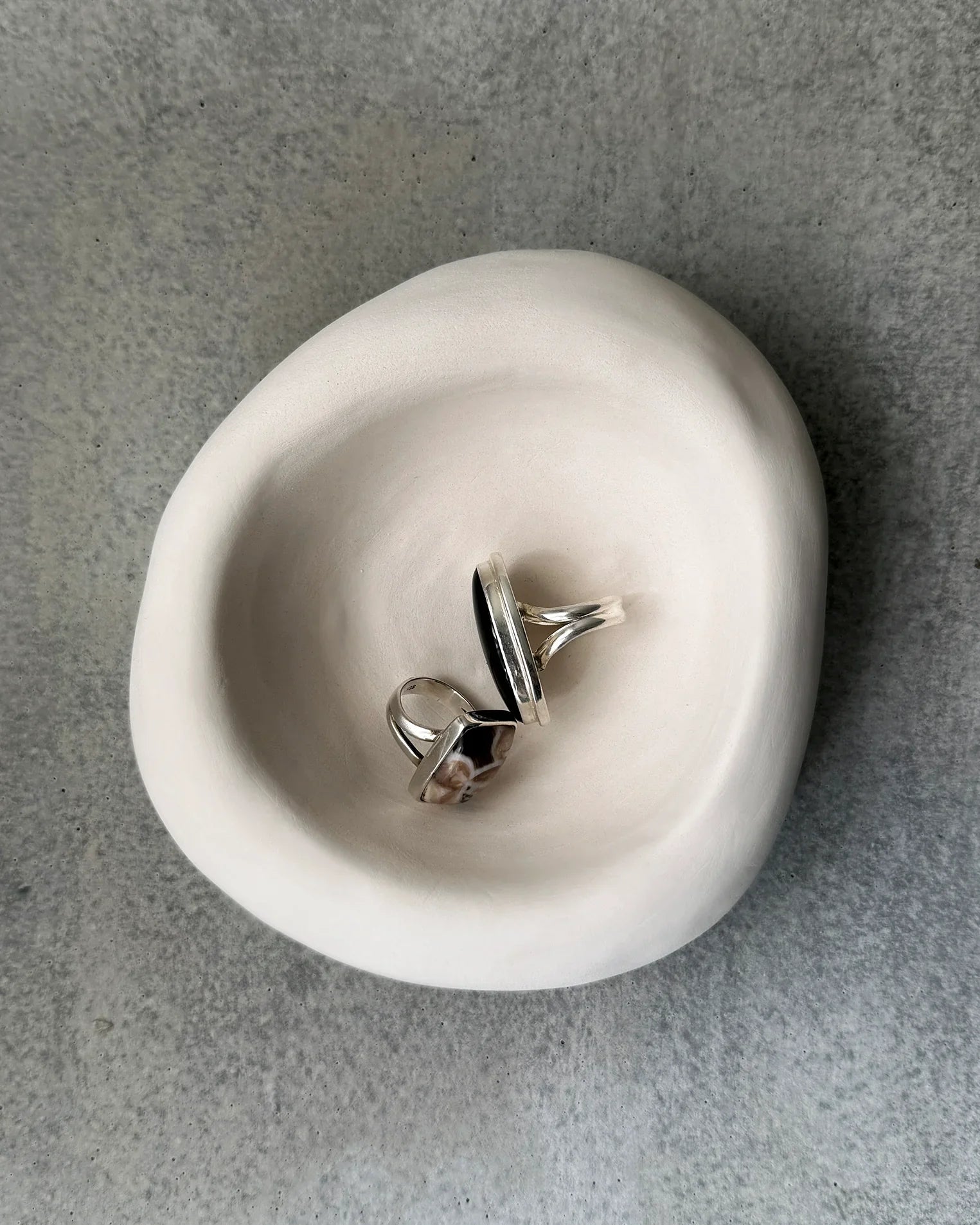Two silver rings with black and patterned stones displayed in a smooth white ceramic bowl on gray stone surface