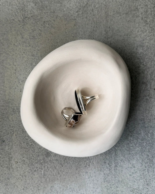 Two silver rings with black and patterned stones displayed in a smooth white ceramic bowl on gray stone surface
