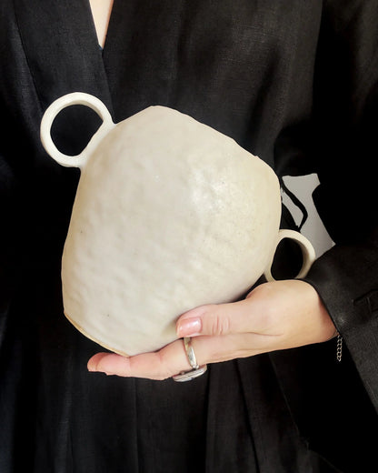 Hand holding a white ceramic vase with two circular handles against a black fabric background