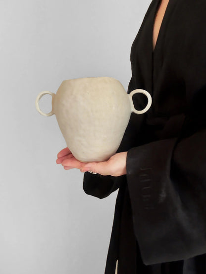 Person in black robe holding a textured white ceramic vase with two round handles