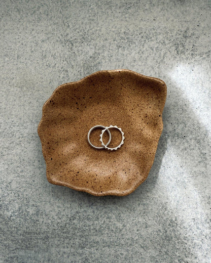 Two silver rings on a textured brown ceramic oyster-shaped dish on a gray surface