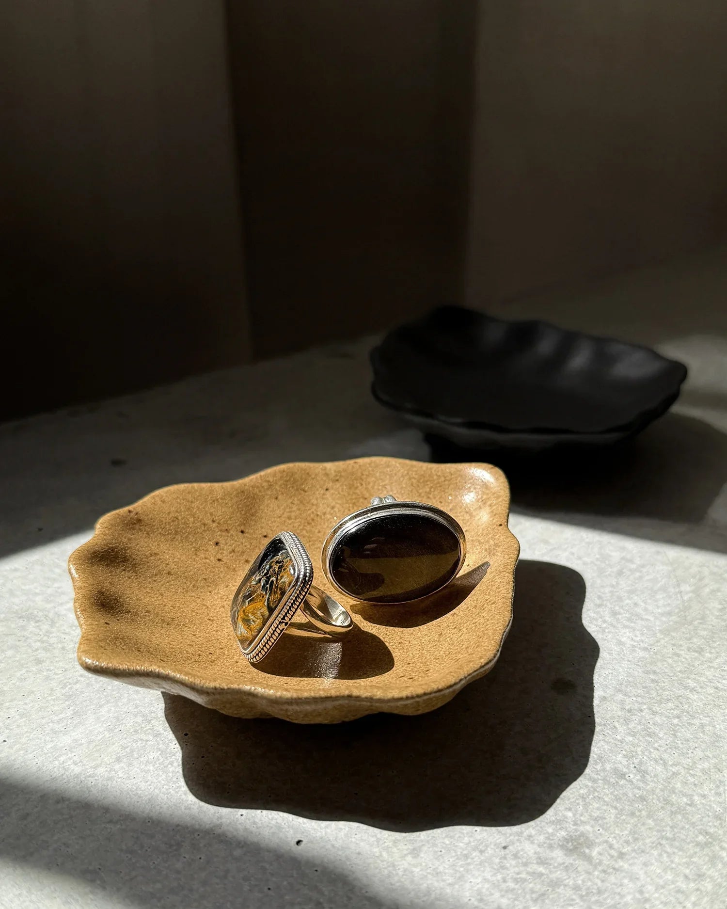 Two silver rings with dark stones on a textured beige ceramic oyster-shaped plate with a black plate in the background