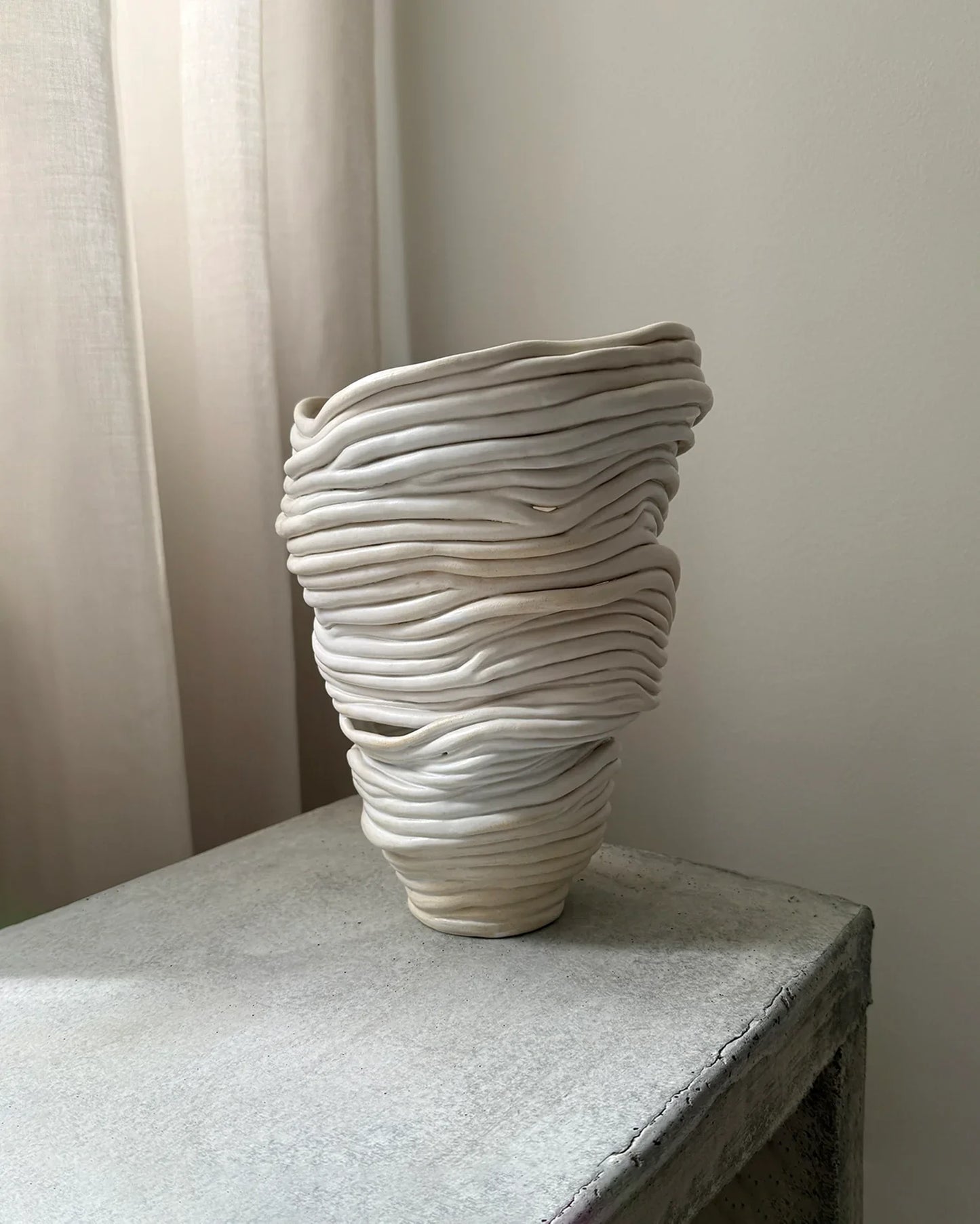 Handcrafted white ceramic vase with layered, wavy design on concrete surface near beige curtains