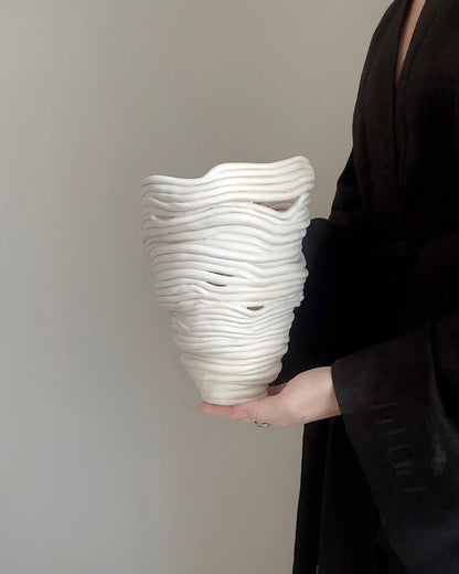Hand holding white ceramic vase with layered, wavy sculptural design against neutral background
