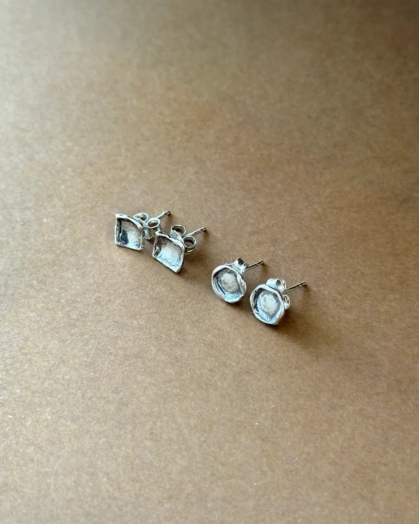 Two pairs of silver stud earrings with square and round shapes on brown surface