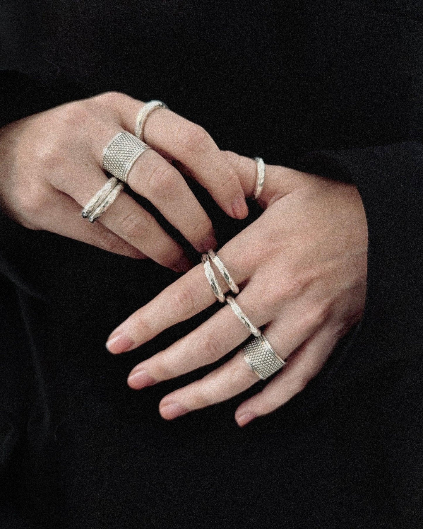 Hands wearing multiple silver rings with textured designs on a black background