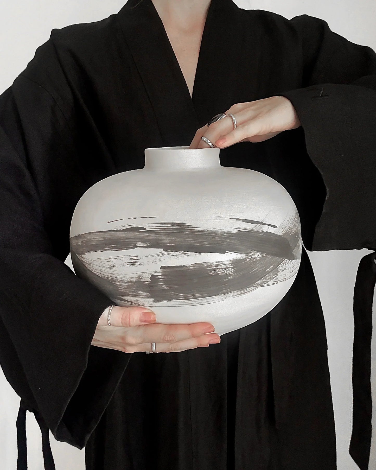 Person in black robe holding white ceramic vase with abstract gray brush strokes